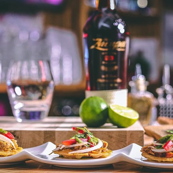 Three small, savory appetizers with shredded meat, onions, and peppers on white plates, with limes and a bottle in the background.
