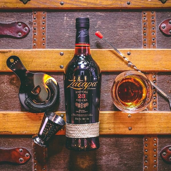 Ron Zacapa Centenario rum bottle, jigger, strainer, and glass on a vintage wooden trunk.
