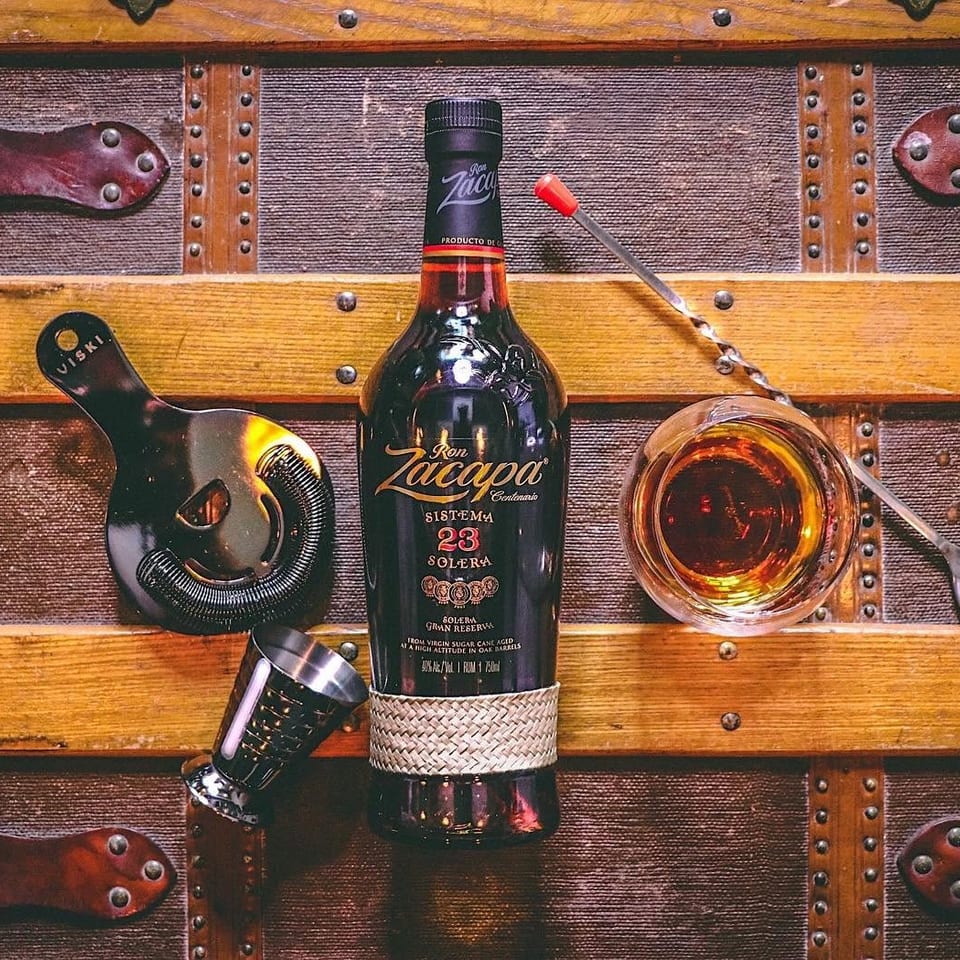 Ron Zacapa Centenario rum bottle, jigger, strainer, and glass on a vintage wooden trunk.