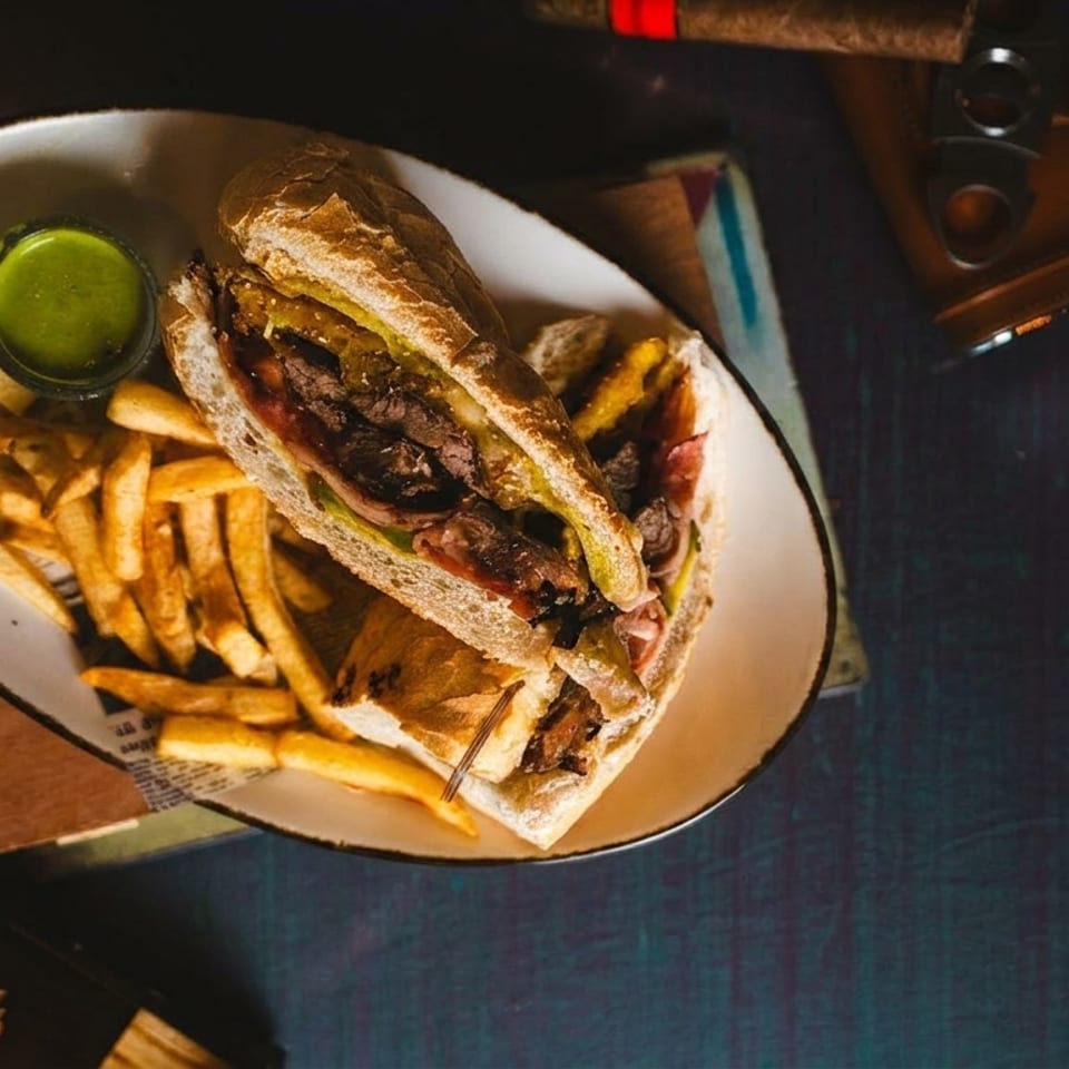 Steak sandwich and fries with green sauce, cigar, and cutter on a dark table.