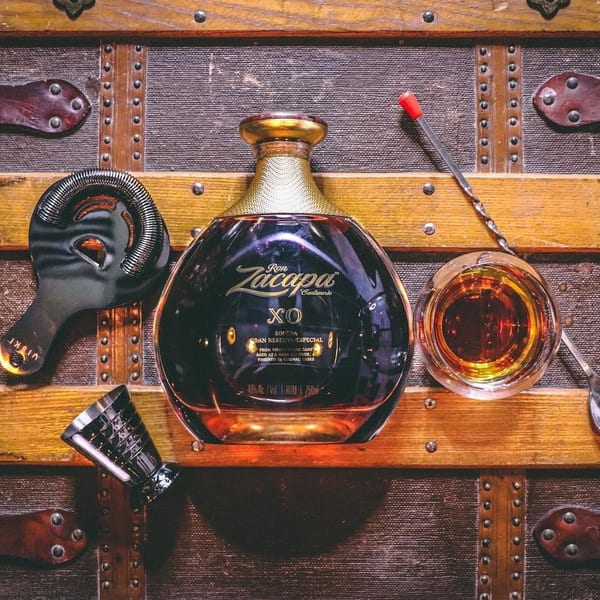 Ron Zacapa XO rum bottle, glass of rum, and bar tools on a vintage wooden trunk.