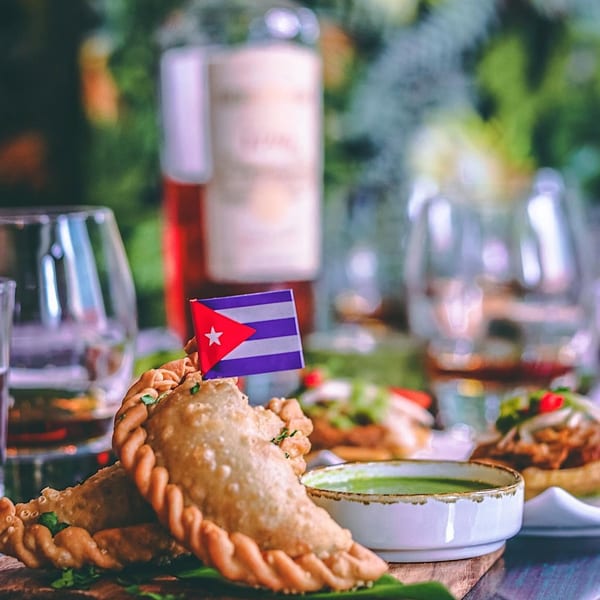 Two golden-brown empanadas with a Cuban flag pick, green sauce, and drinks on a wooden board.